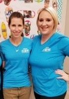 Two women in matching blue shirts smile, standing indoors. The background includes various colorful objects, possibly decorations.