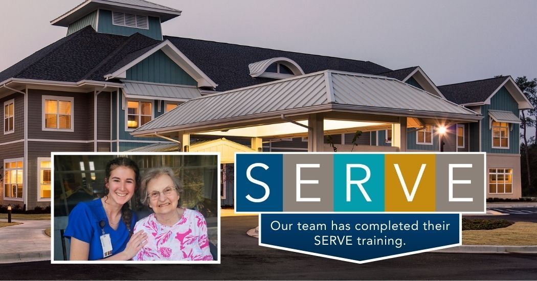 Two nurses are standing next to each other in front of a large house with a sign that says serve