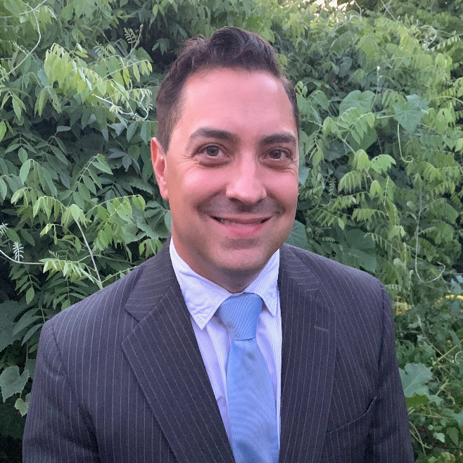 A man in a suit and tie is smiling in front of a bush.