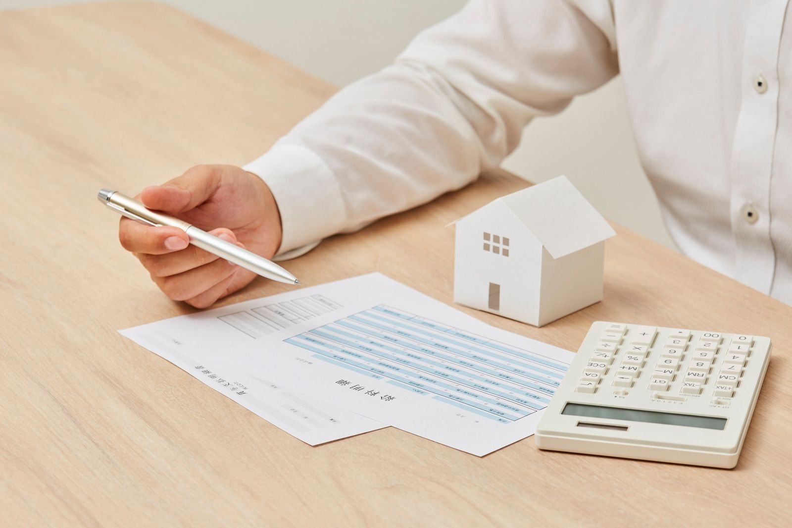 Person with pen reviewing paperwork, small house model, and calculator on a wooden desk.