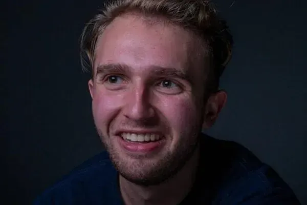 Man with blonde hair smiles, looking off-camera. Dark background, wearing a dark blue shirt.