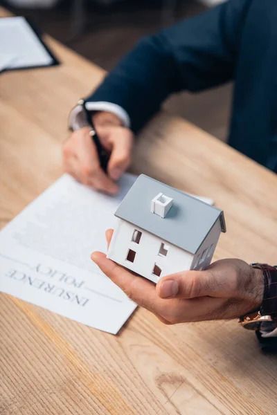 Person holding a miniature house, signing an insurance policy document on a wooden desk.