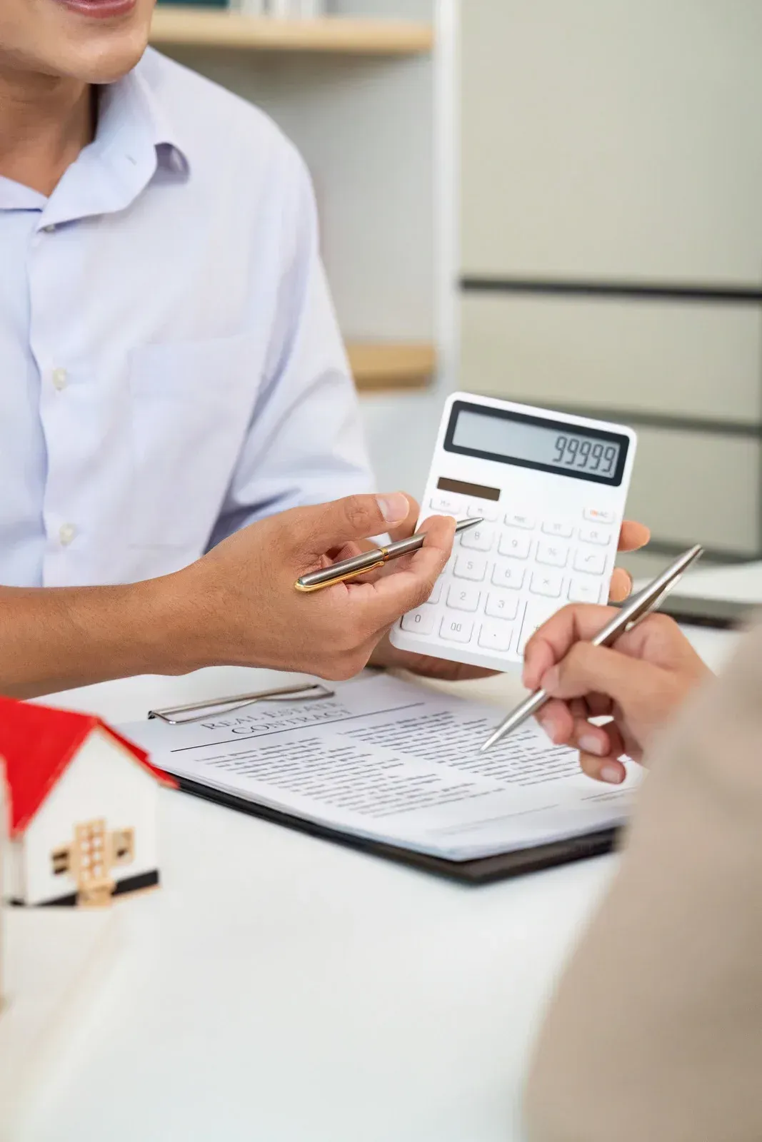 Hands pointing at a calculator and paperwork. A small house model sits on the table.