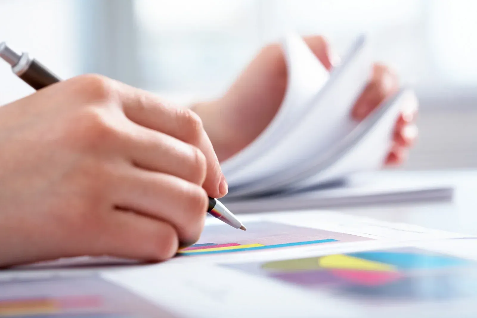 Person writing on a document with colorful charts, flipping pages with the other hand.