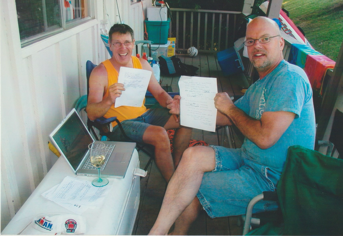 Two men sit on a porch holding papers and a laptop