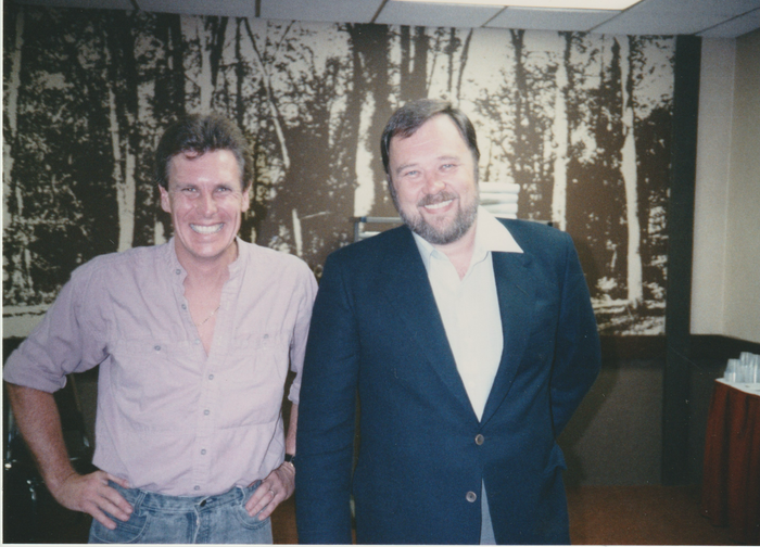 Two men are posing for a picture in front of a wall with trees on it