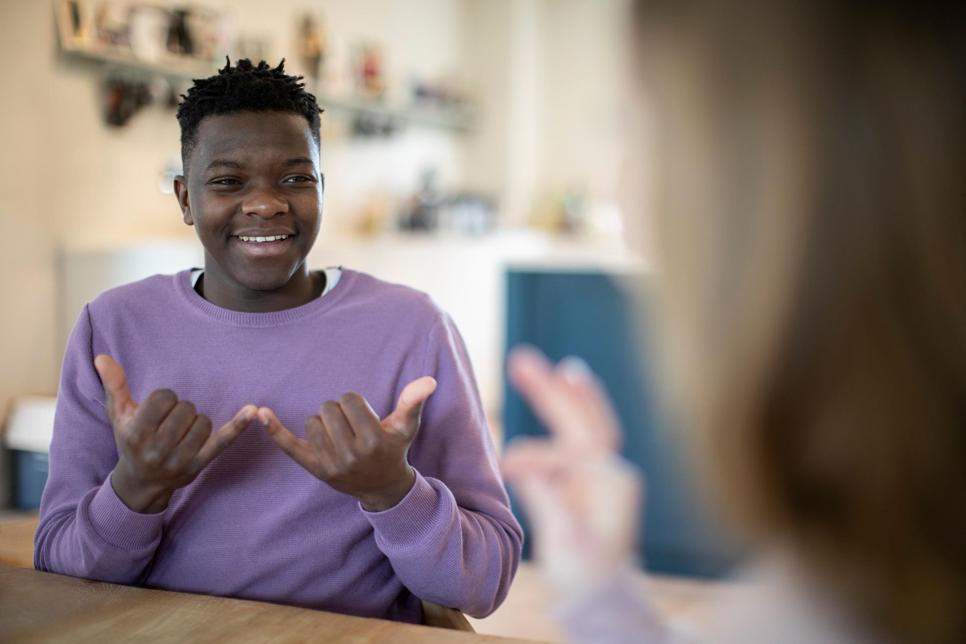 Young Black man signing with hands, smiling, interacting with someone out of frame. Indoors, natural light.