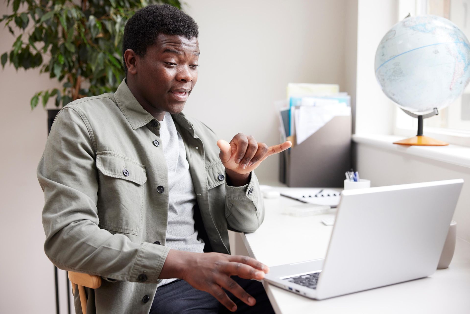 Man in a gray shirt signing in front of a laptop at a desk near a window with a globe.