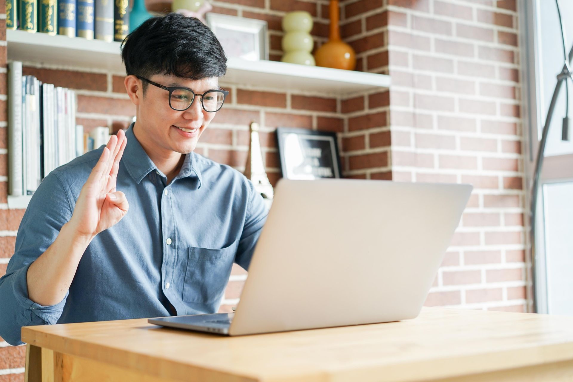 Man with glasses and blue shirt waves at laptop; seated at a desk in a brick-walled room.