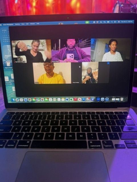 Laptop screen displaying a video call with five people. People wave and gesture. Keyboard visible.