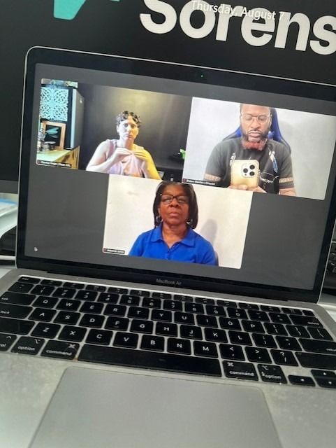 Laptop screen shows three people in a video call. Woman signing, man holding phone, another woman wearing blue.