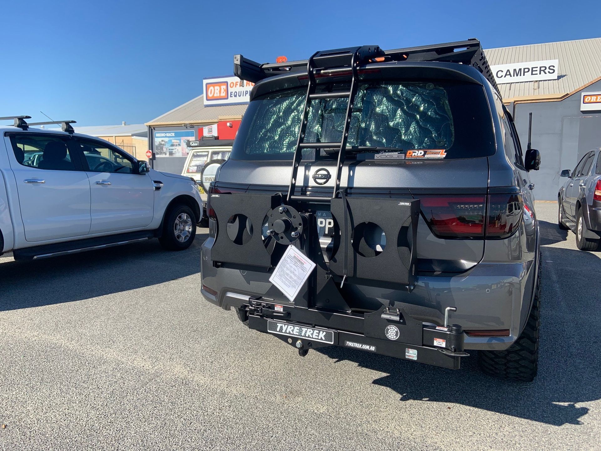 A car with a ladder and tyre rack attached to the back of it is parked in a carpark.