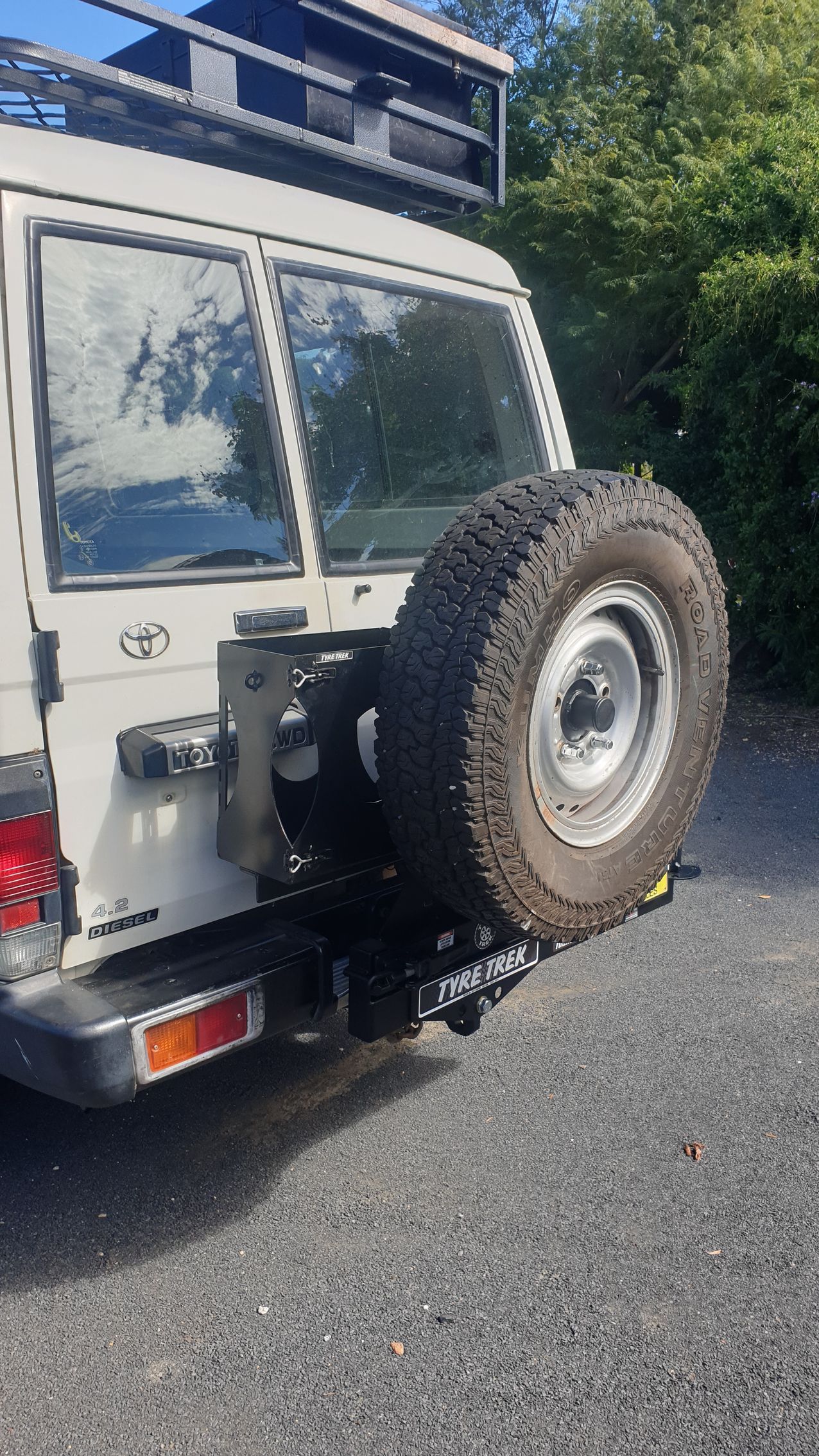 A white jeep with a spare tyre attached to the back is parked in a gravel carpark.