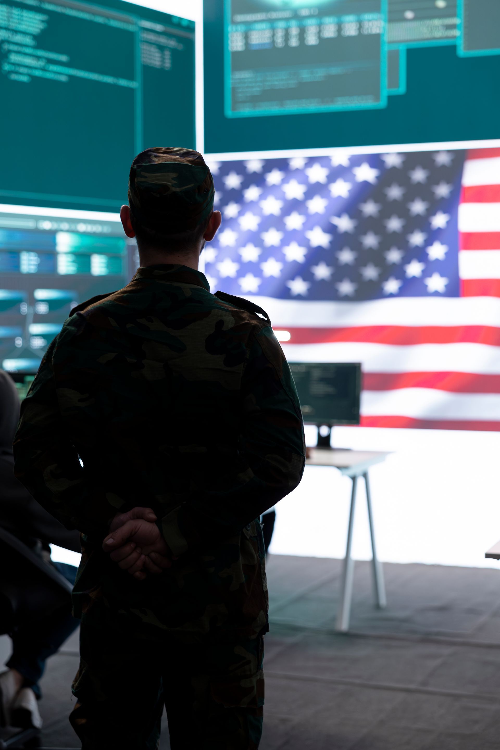 A soldier is standing in front of a large screen with an american flag in the background.