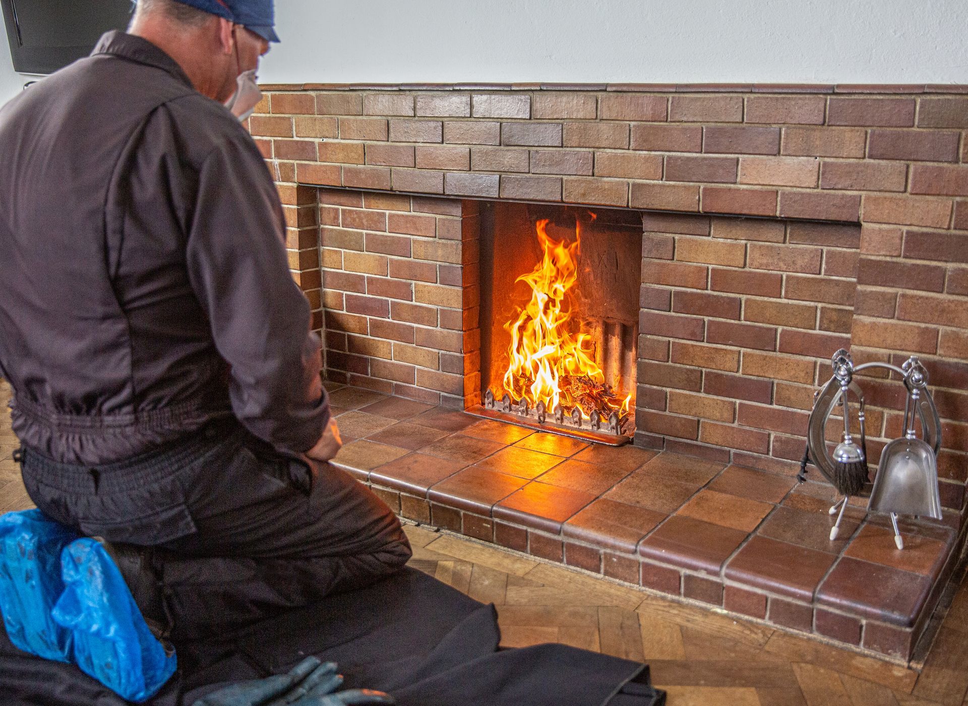 Person kneeling by a brick fireplace, watching flames. Dark clothing, blue hat, tools nearby.