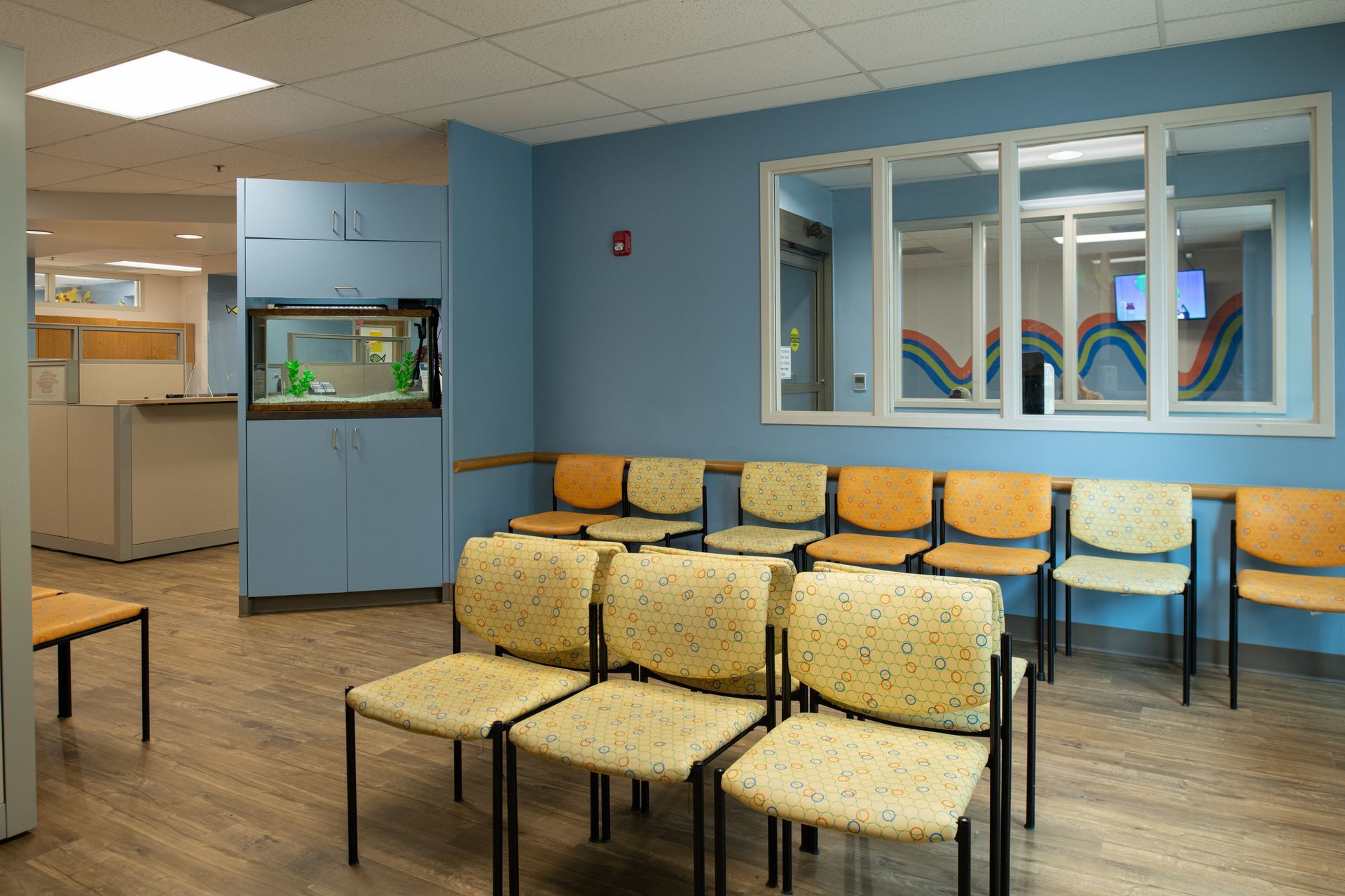 A light blue children's waiting room with chairs, a fish tank, and a window with a rainbow mural.