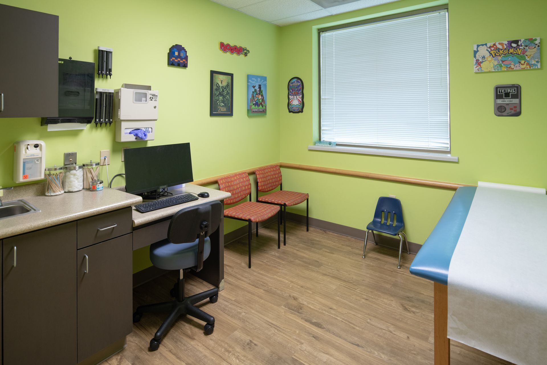 Doctor's office, green walls, desk with computer, chairs, examination table, and artwork for children.