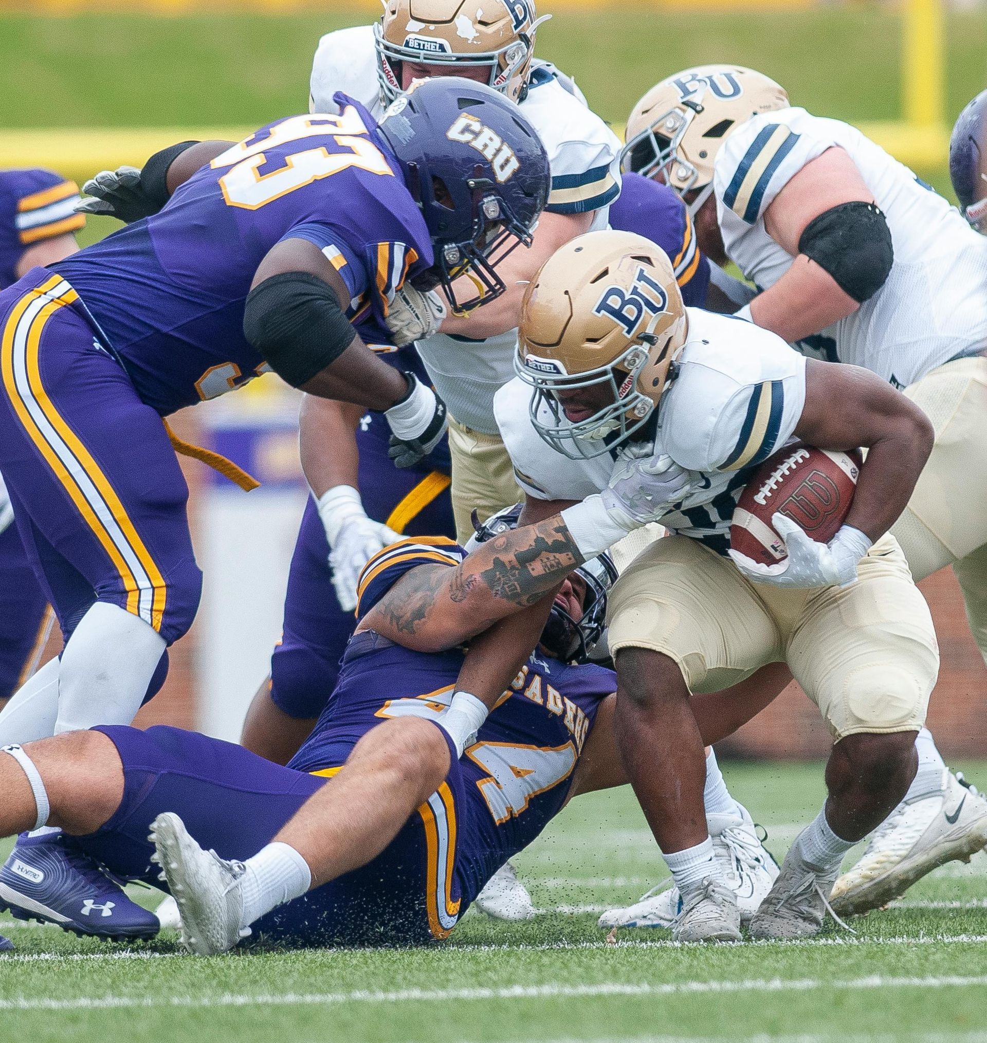 Football players in purple and gold uniforms tackle a player in a white and gold uniform on a grassy field.