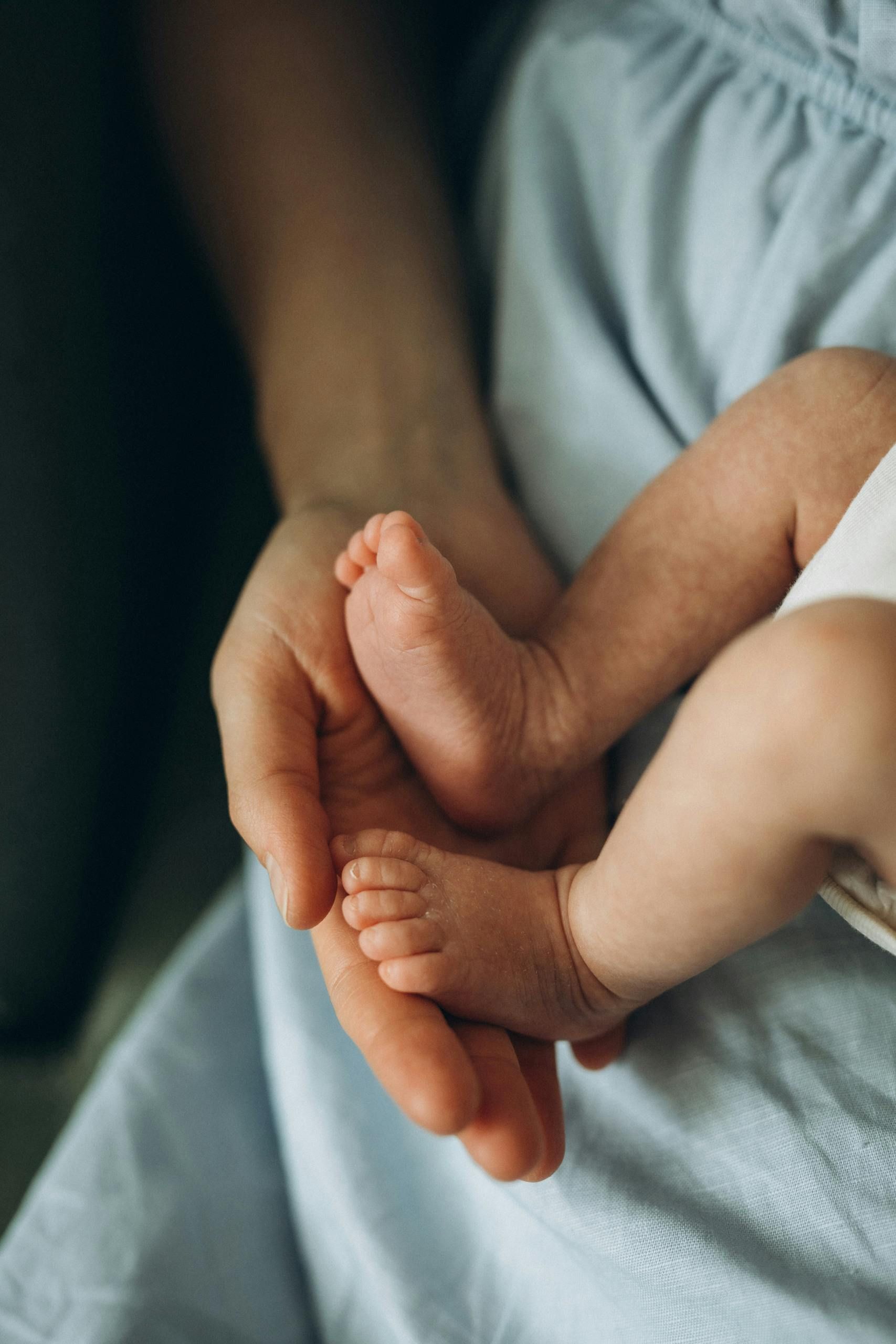 Person's hands cradling a newborn baby's tiny feet, set against a blue dress.
