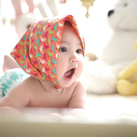 Baby with open mouth wearing an orange patterned head covering; lying on its stomach, surrounded by toys.