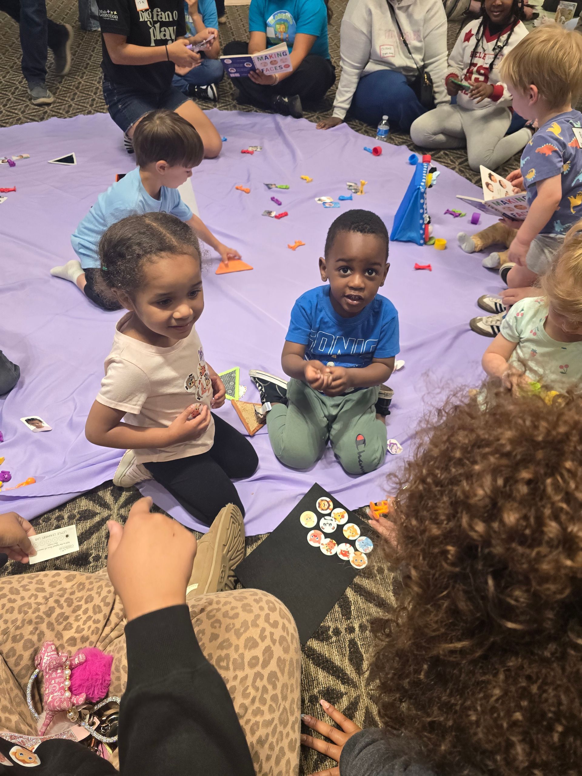 Children sitting on a purple mat during a group activity, with scattered craft pieces and adults nearby