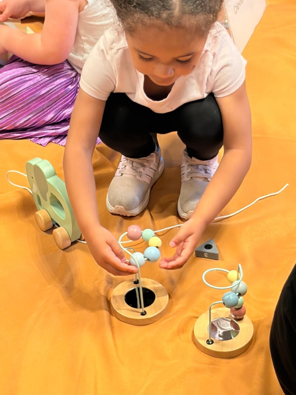 Child bending over a wooden bead maze toy on an orange mat, hands moving pastel beads.