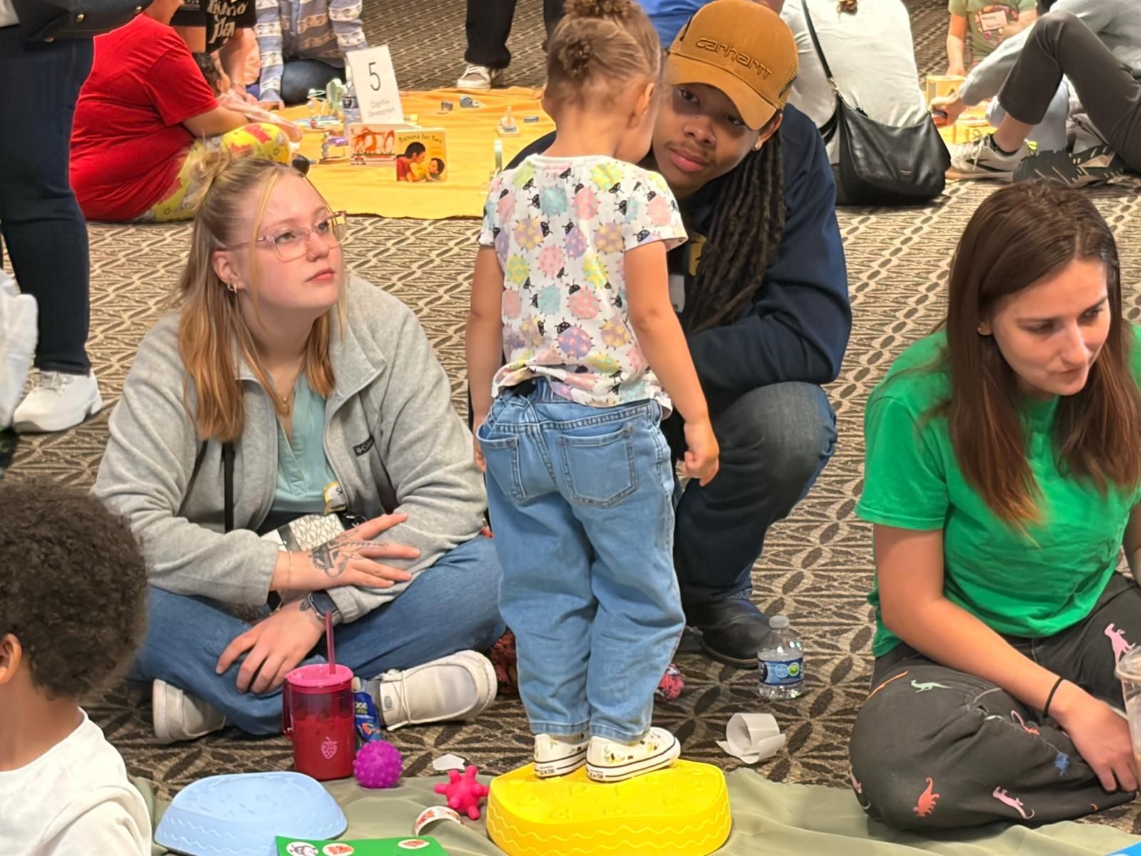 People sitting on the floor in a casual indoor gathering, with colorful toys and cups nearby.
