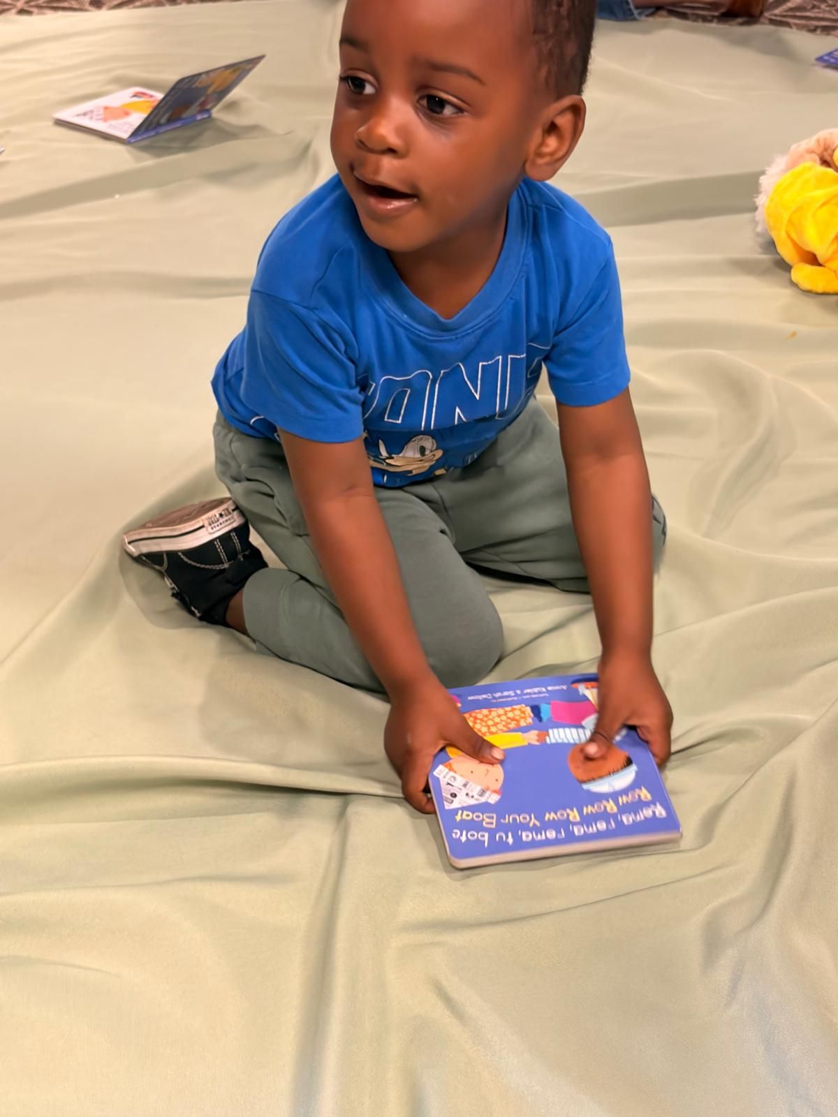Child sitting on a mat holding a purple book, looking up in a classroom-like setting.