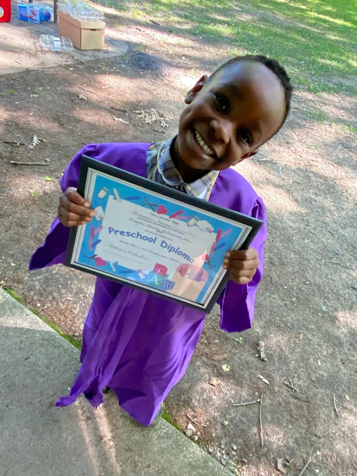 Smiling child in purple outfit holding a framed certificate outdoors on a dirt path.