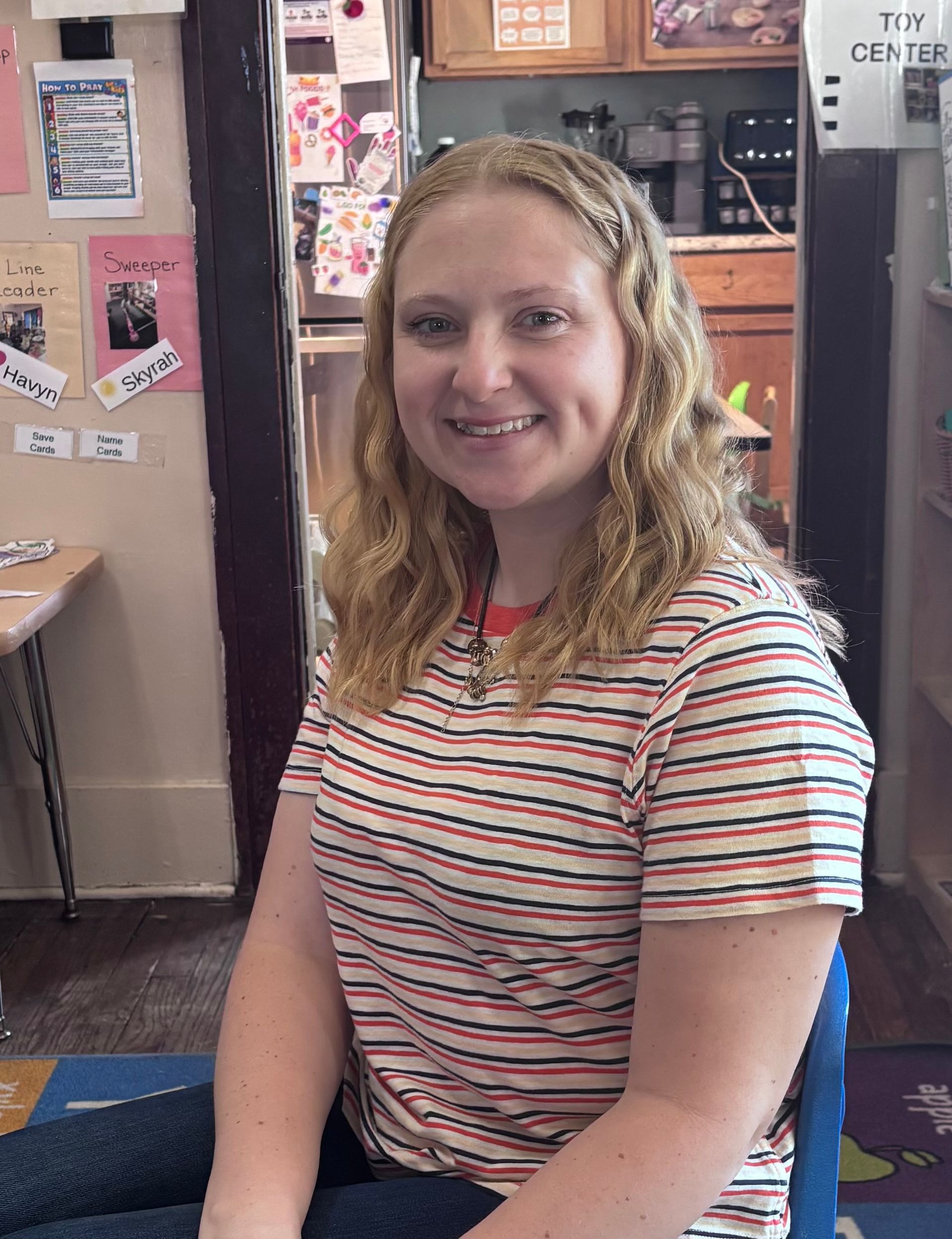 Smiling woman in a striped shirt seated indoors against a bulletin-board background