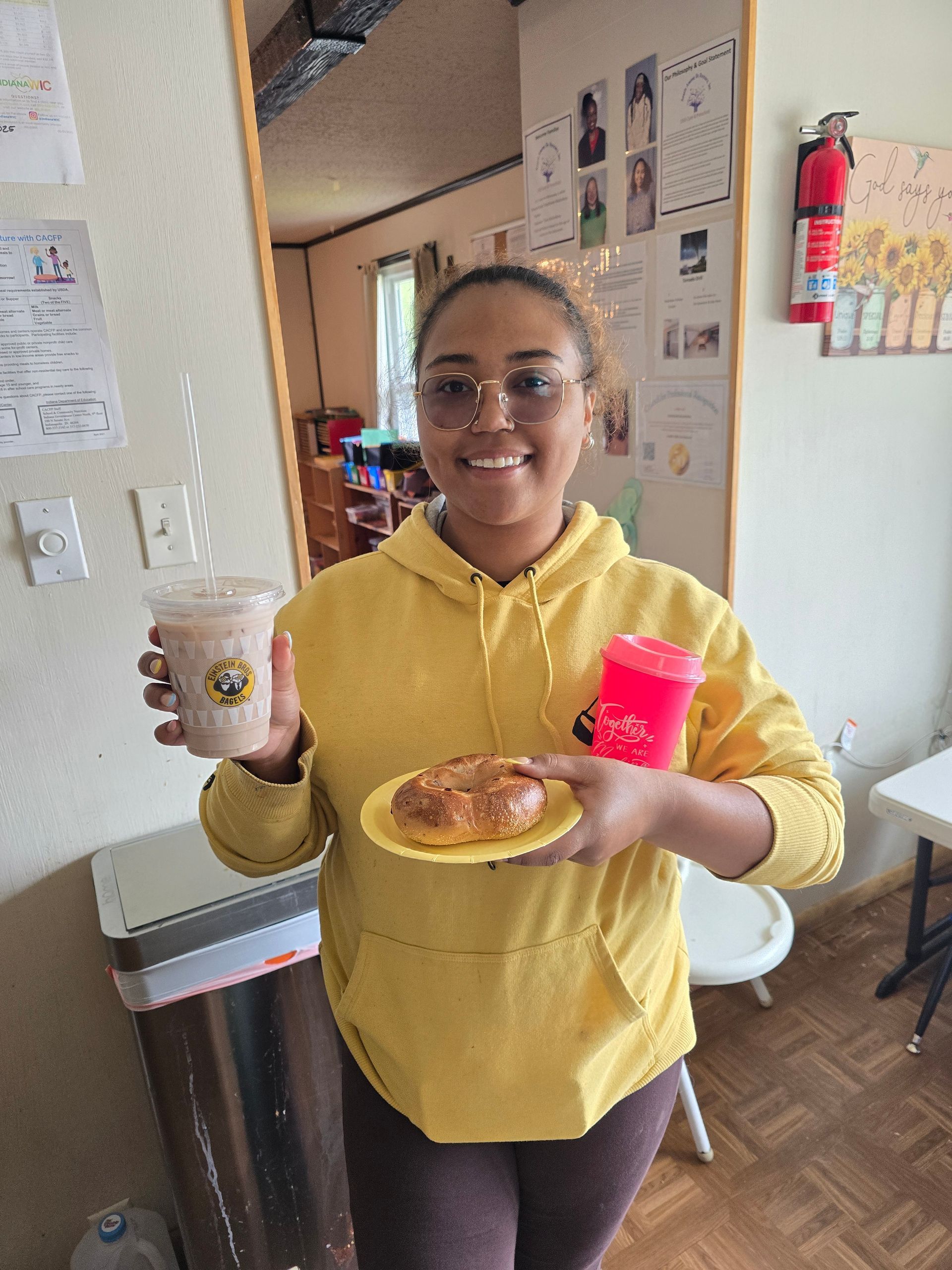 Person in a yellow hoodie holding a drink, donut, and pink cup in a home kitchen.