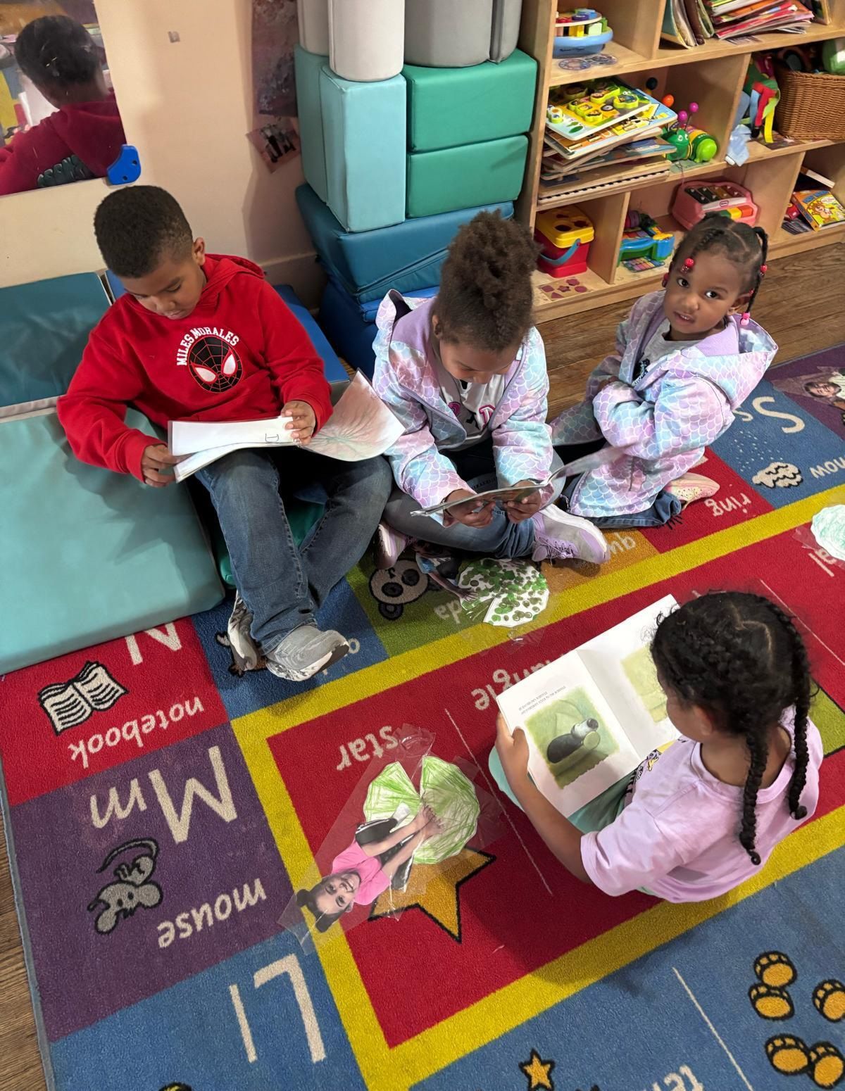 Children sitting on a colorful classroom rug, reading books together near shelves and bins.