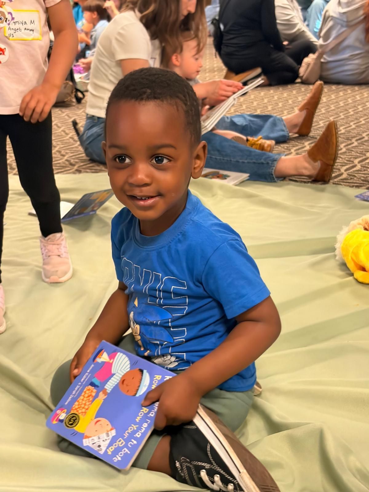 Child in blue shirt holding a book while sitting on a mat among other children.