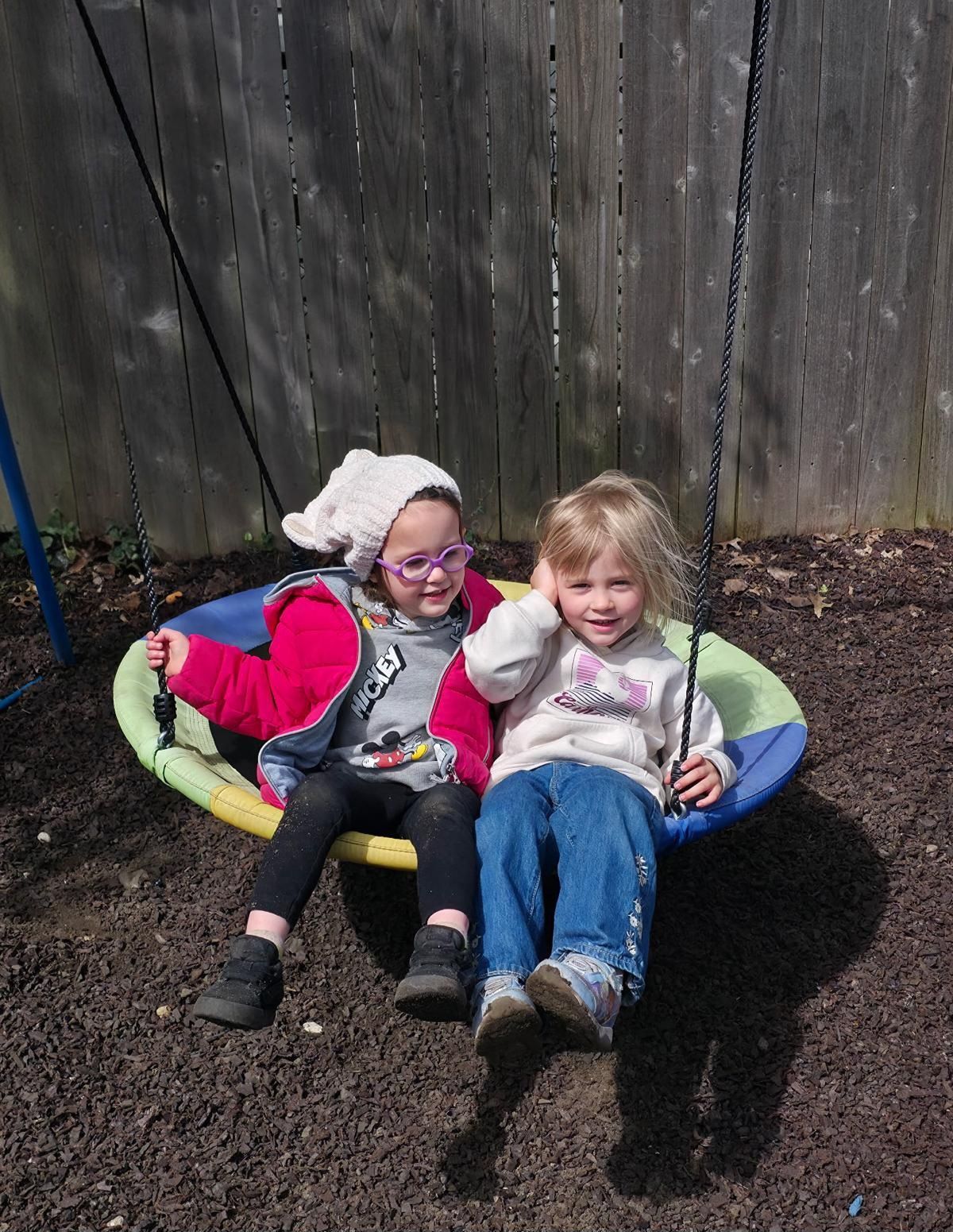 Two children sit together on a colorful swing seat outdoors, smiling by a wooden fence.