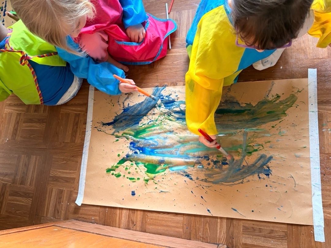 Three children in a bright classroom playing with colorful handbells on a white table.