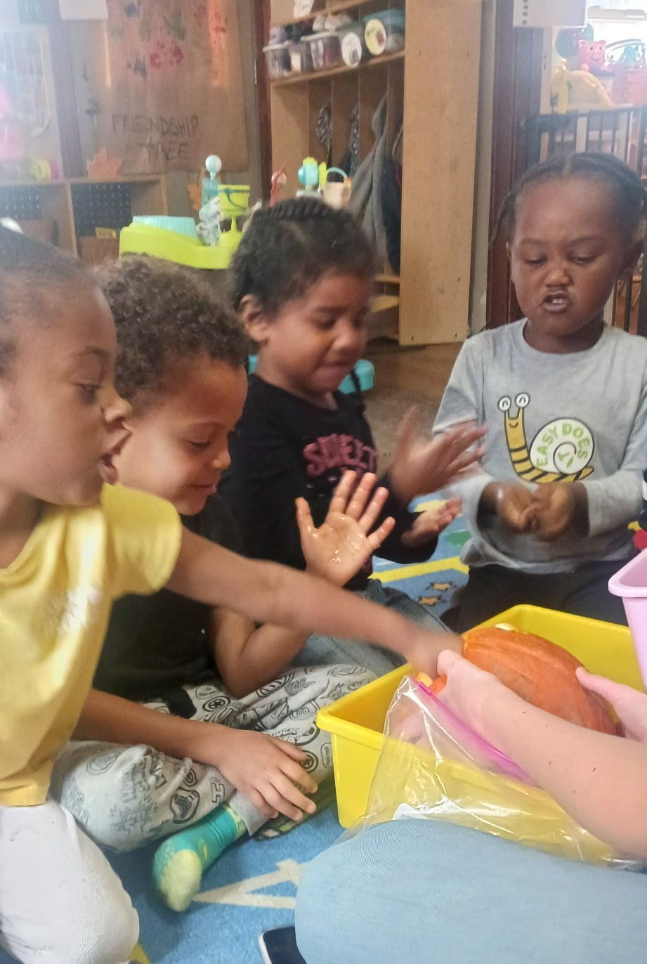 Four children sitting around a green table in a classroom with a chalkboard, coloring and drawing together.