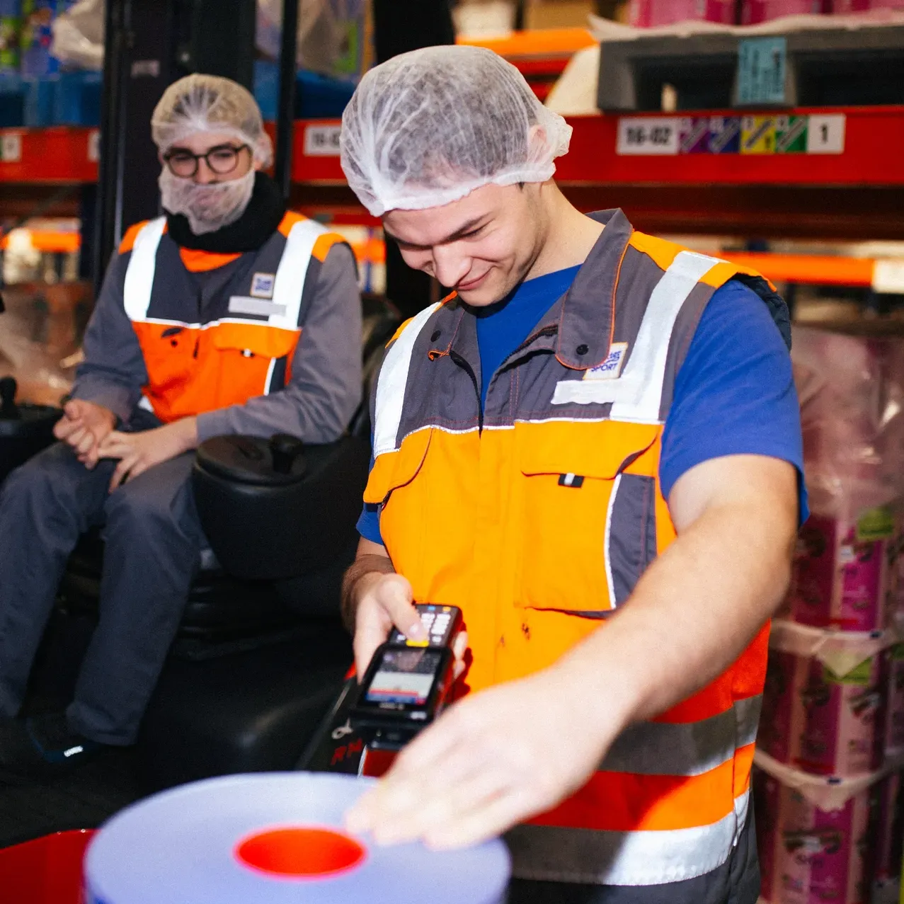 Two men wearing safety vests and hair nets are working in a warehouse