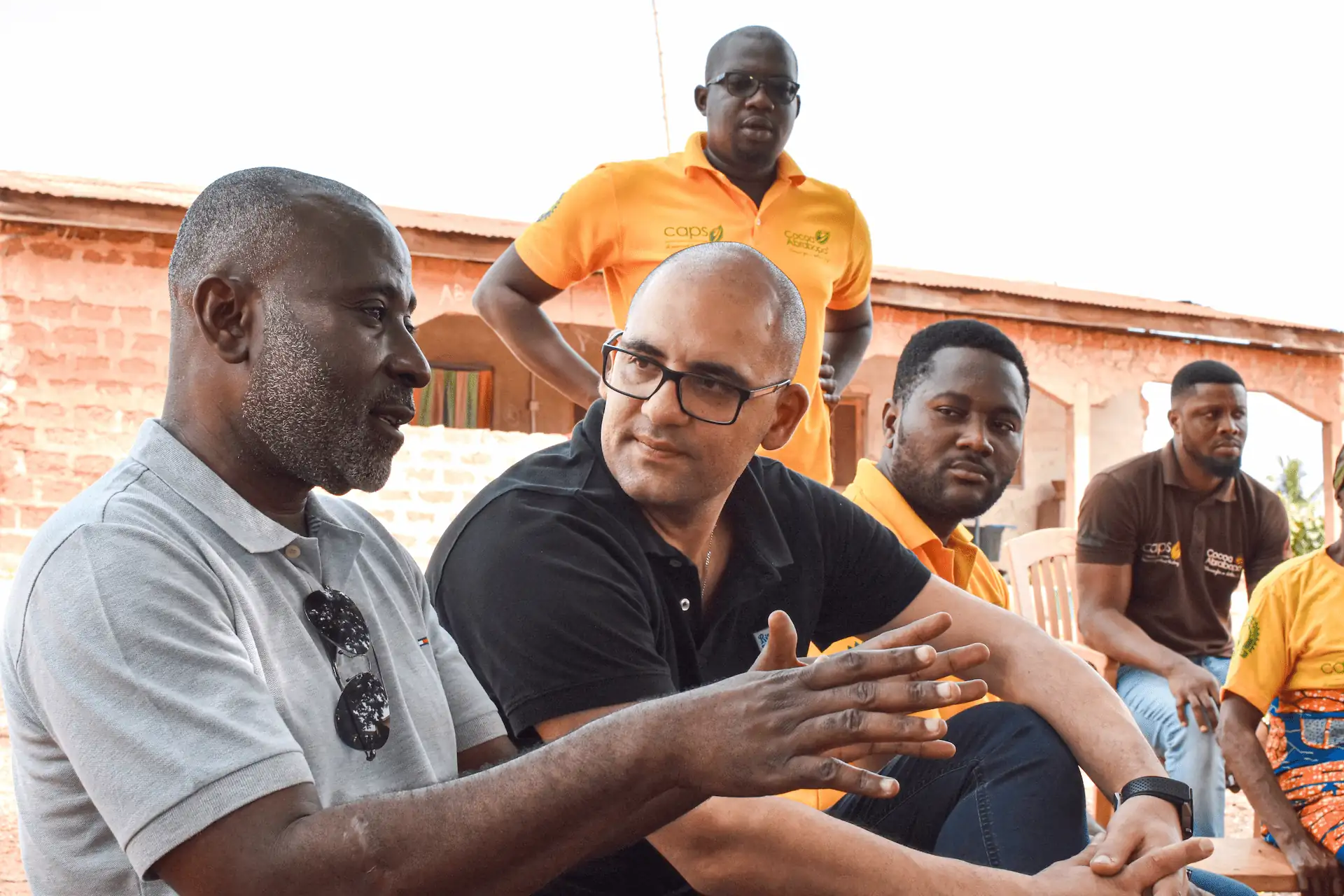 Group of men in discussion outdoors; one gestures with hands.