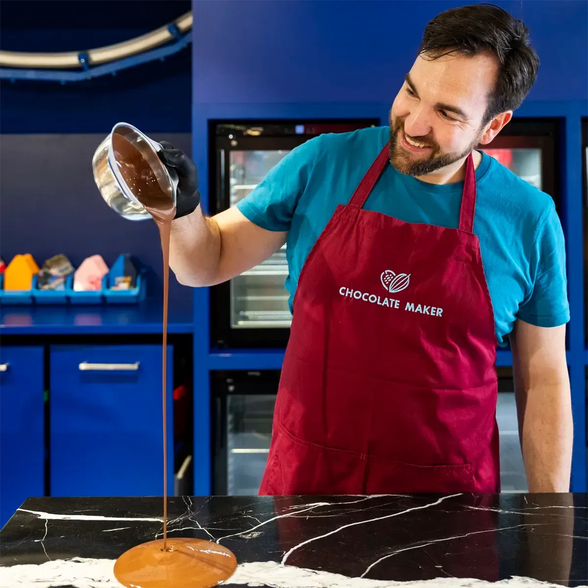 Ein Chocolatier in blauem Hemd und bordeauxroter Schürze gießt geschmolzene Schokolade auf eine schwarze Marmorarbeitsplatte.