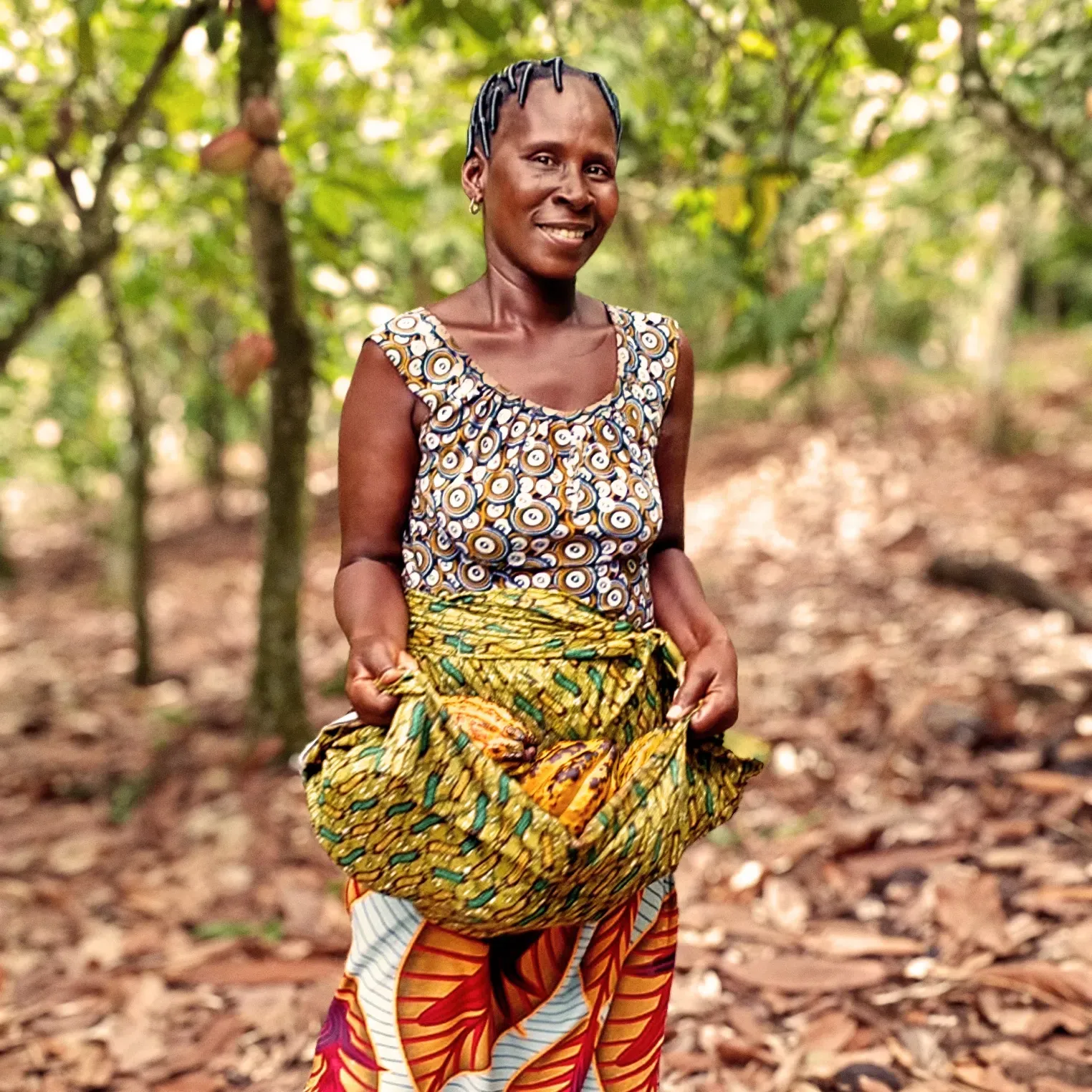 Woman in a cocoa farm holding a bag of yellow pods, smiling, trees in the background with fallen leaves.