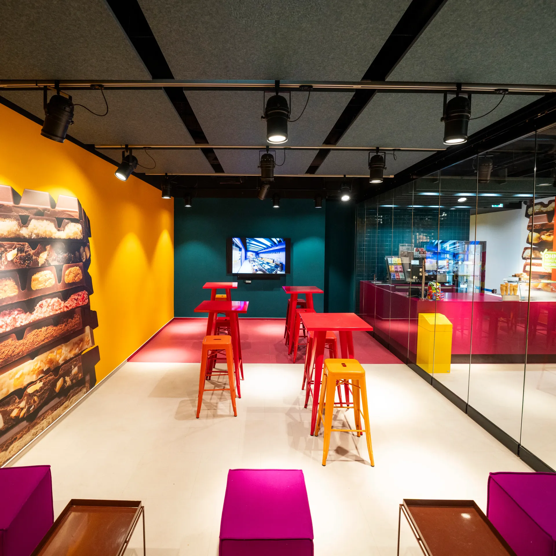 Ein moderner, farbenfroher Loungebereich mit hohen Bartischen, leuchtend orangefarbenen Hockern, einer schokoladenbraunen Wandgrafik und einem Fernsehbildschirm.