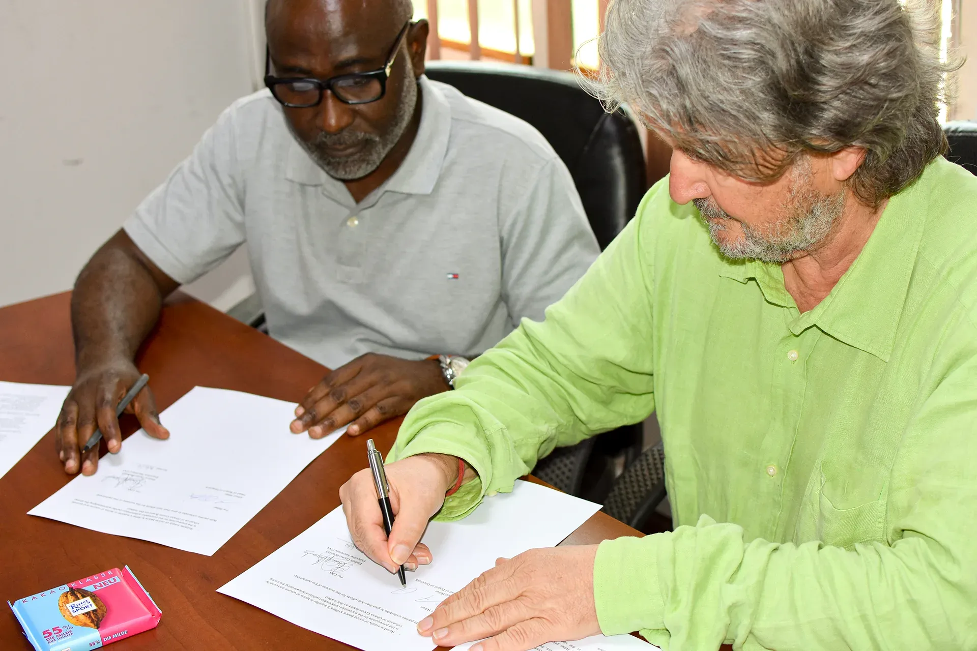 Two men signing documents at a table. One wears glasses, the other a green shirt.