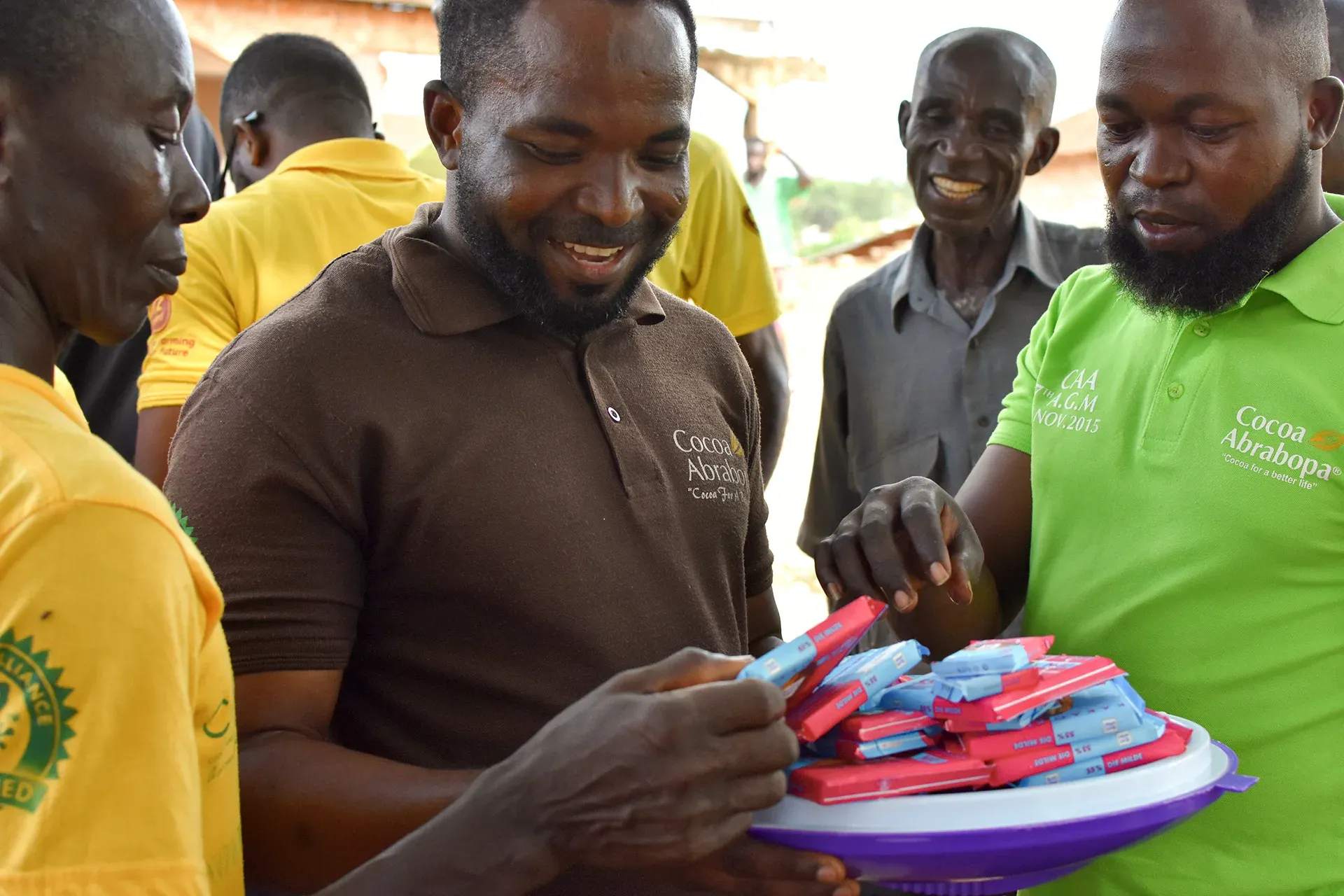 Men in a village are smiling and holding colorful items, possibly distributing them in a community setting.