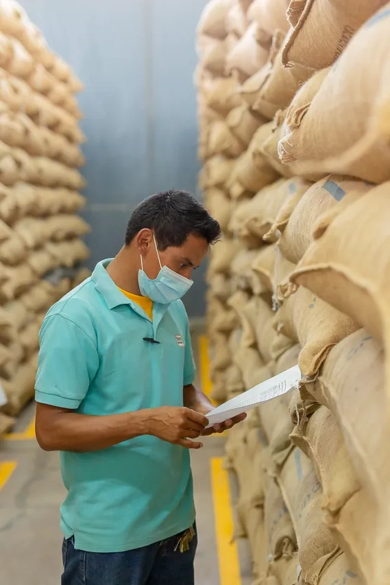 Man in mask inspecting documents, surrounded by stacked burlap sacks in a warehouse.