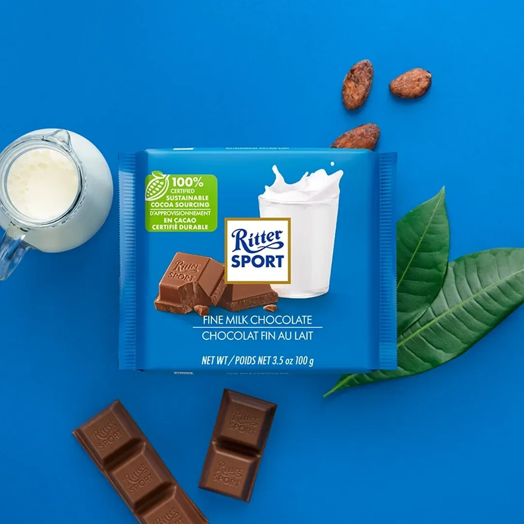 Ritter Sport milk chocolate bar with milk splash graphic, cocoa beans, milk jug, and blue background.