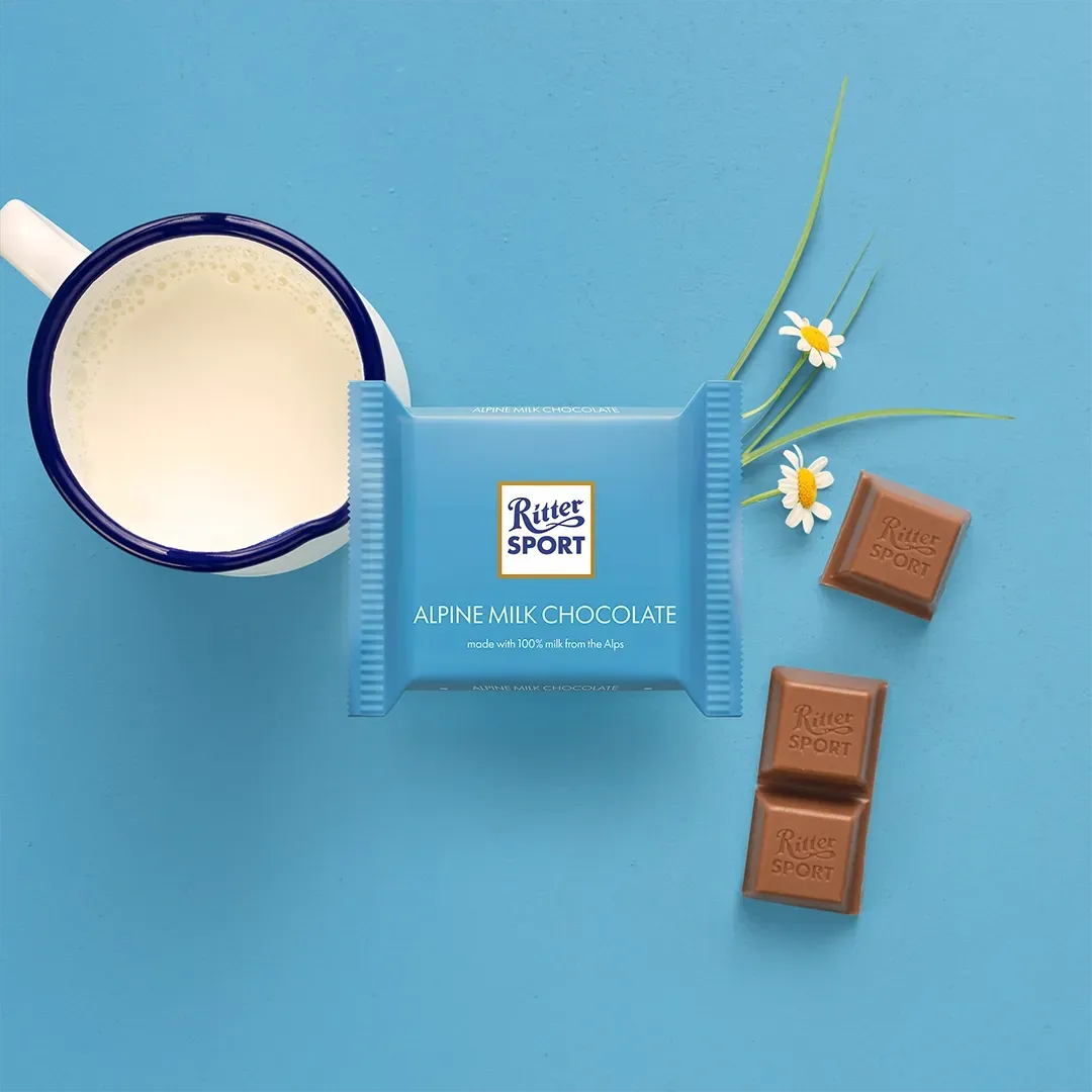 Ritter Sport almond chocolate bar on blue, with mug of milk and flowers.