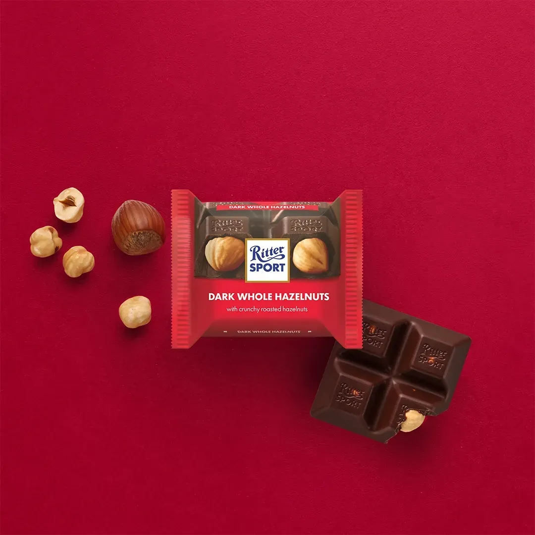Ritter Sport dark whole hazelnut chocolate bar on a red background with loose hazelnuts and a broken piece of chocolate.