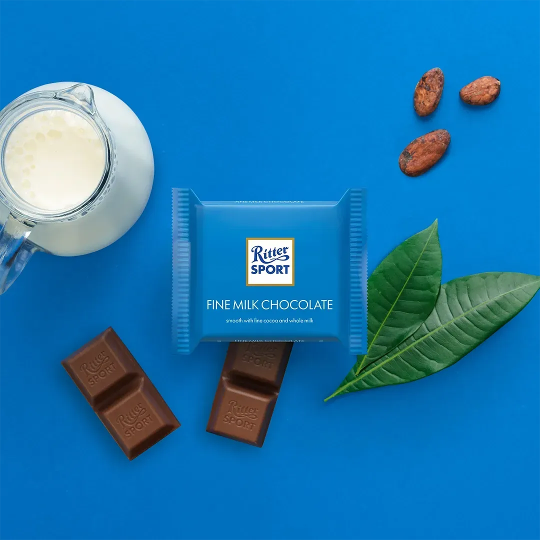 Blue Ritter Sport chocolate bar with cocoa beans, leaves, chocolate pieces, and a milk pitcher on a blue surface.