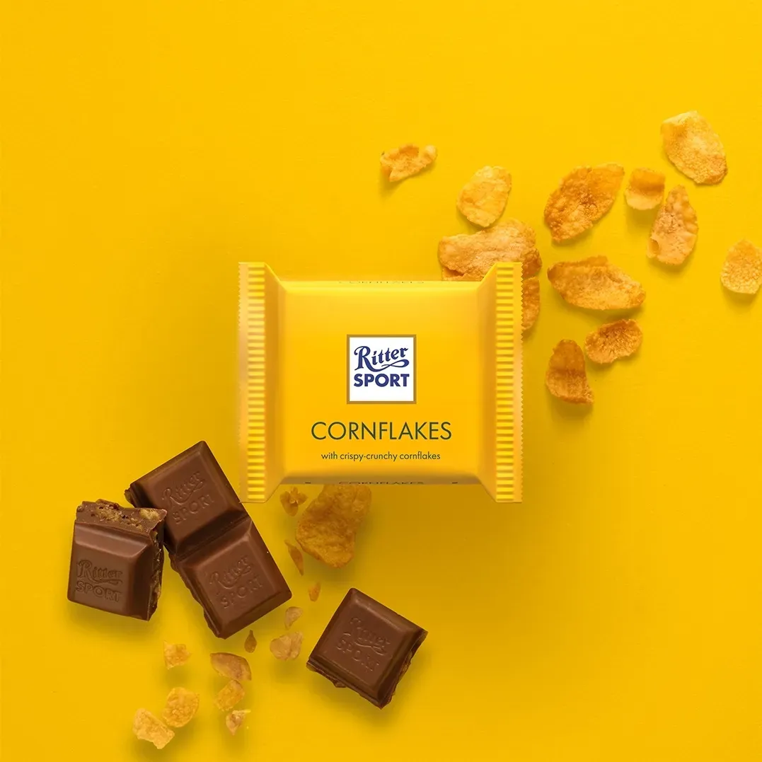Ritter Sport Cornflakes chocolate bar on yellow background with cornflakes and chocolate pieces.