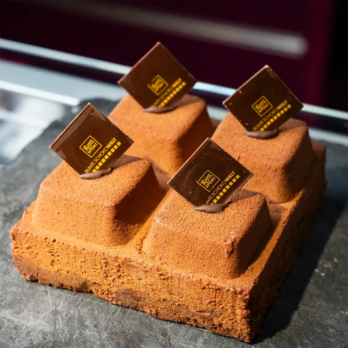 Ein quadratisches Schokoladendessert mit einer strukturierten, braunen Oberfläche und vier erhabenen Segmenten, die jeweils mit einem Ritter Sport Quadrat verziert sind.